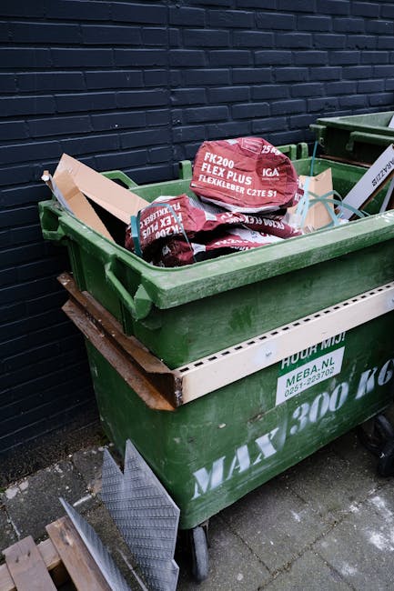 A large green wheeled rubbish bin filled with red and black bags of garden or building waste, branded with product labels, sits on a paved sidewalk against a dark blue brick wall. The bin is partially
