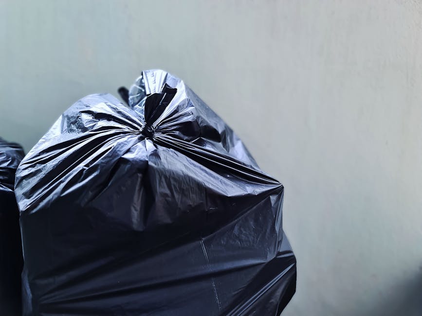 A large green wheeled rubbish bin filled with red and black bags of garden or building waste, branded with product labels, sits on a paved sidewalk against a dark blue brick wall. The bin is partially