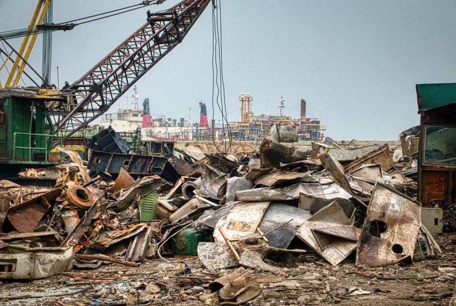 A large heap of assorted metallic debris consisting of rusted and weathered sheet metal, old appliances, and various scrap parts is piled in an industrial yard. The materials show a mix of textures, i